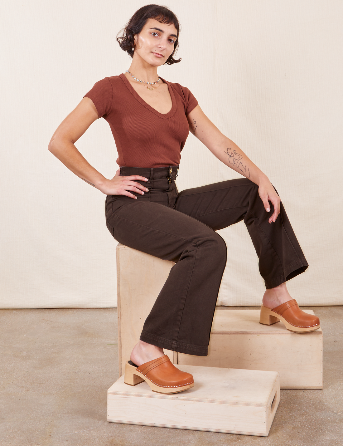 Soraya sitting on a wooden crate wearing Western Pants in Espresso Brown paired with fudgesicle brown V-Neck Tee