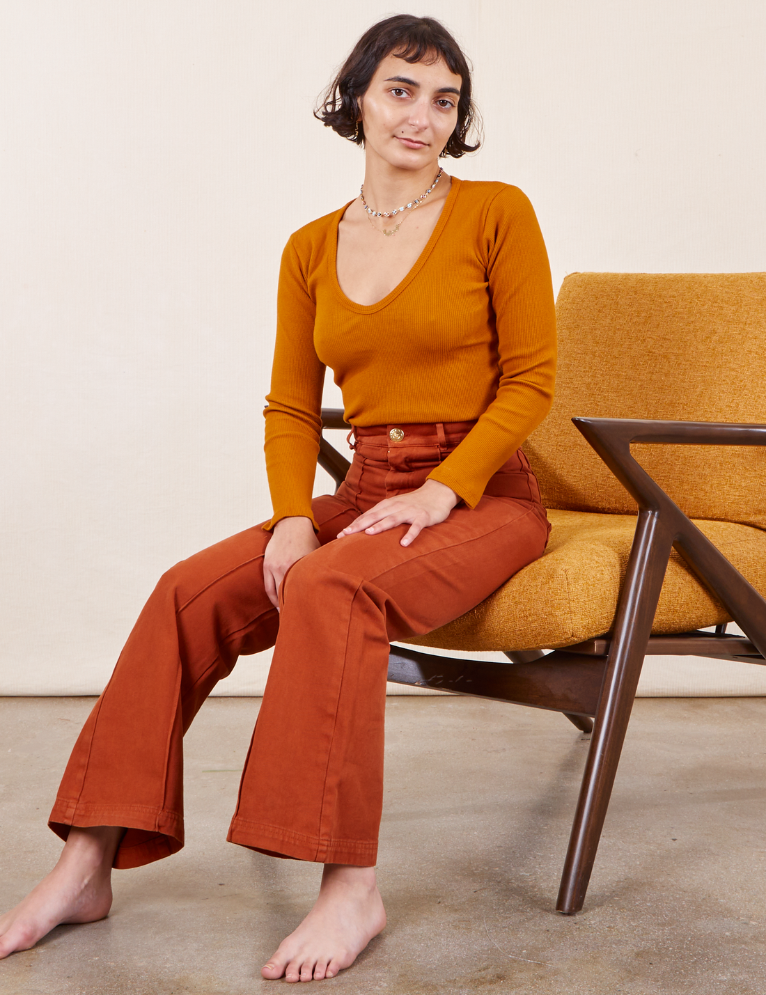 Soraya is sitting in an orange upholstered chair. She is wearing Western Pants in Burnt Terracotta paired with a burnt orange Long Sleeve V-Neck Tee