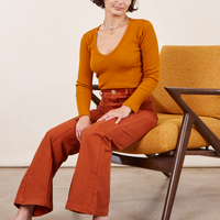 Soraya is sitting in an orange upholstered chair. She is wearing Western Pants in Burnt Terracotta paired with a burnt orange Long Sleeve V-Neck Tee
