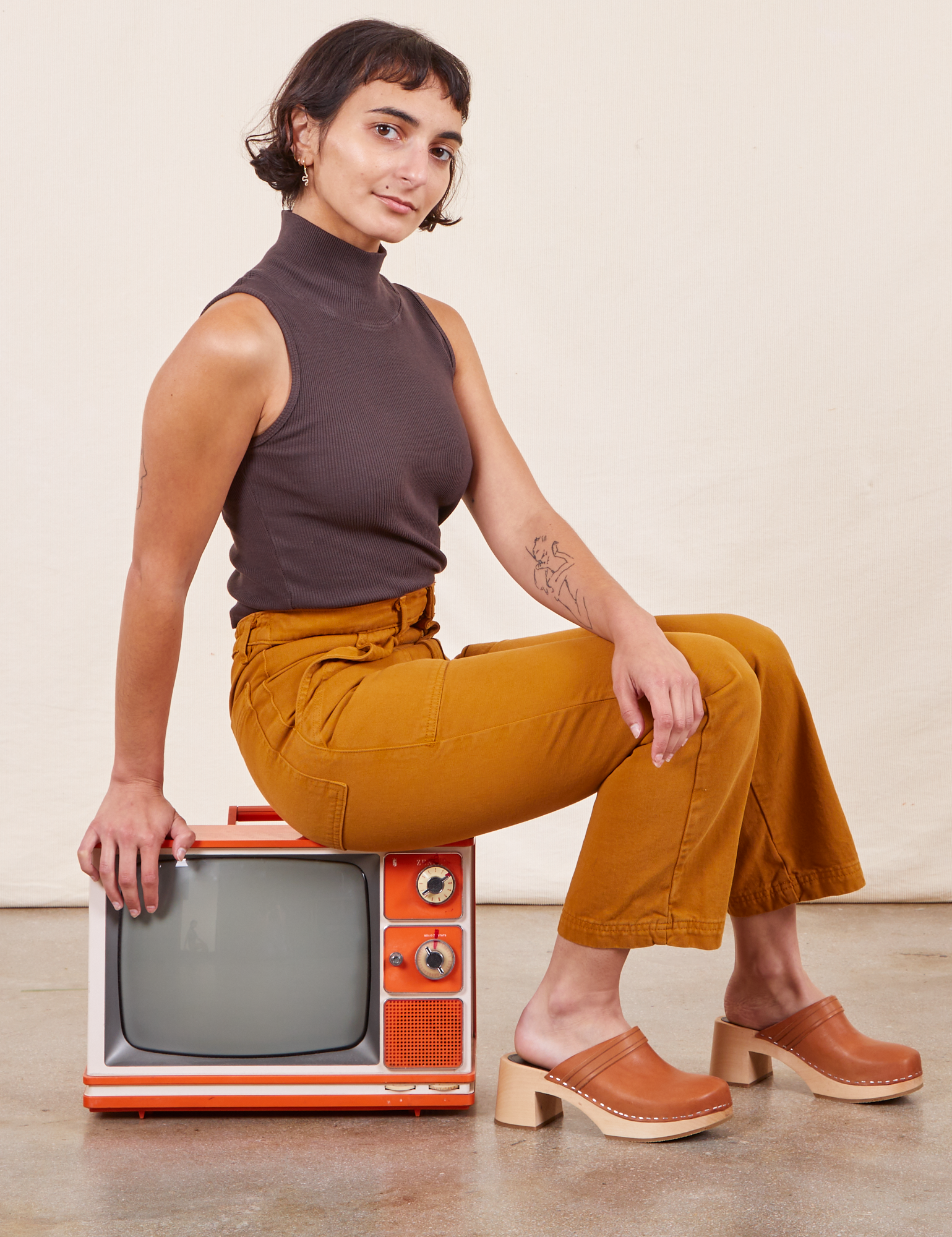 Soraya is sitting sideways on a small orange vintage tv wearing Work Pants in Spicy Mustard and espresso brown Sleeveless Turtleneck