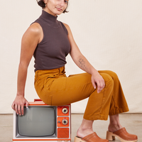 Soraya is sitting sideways on a small orange vintage tv wearing Work Pants in Spicy Mustard and espresso brown Sleeveless Turtleneck
