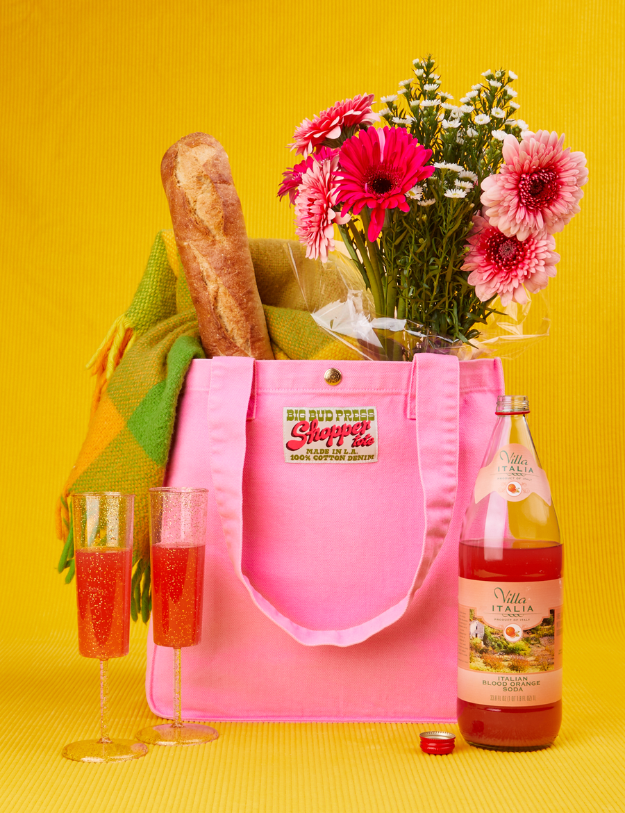 Shopper Tote Bag in Bubblegum Pink with baguette, flowers and scarf inside. Two glasses and a bottle of blood orange soda in front of the bag.