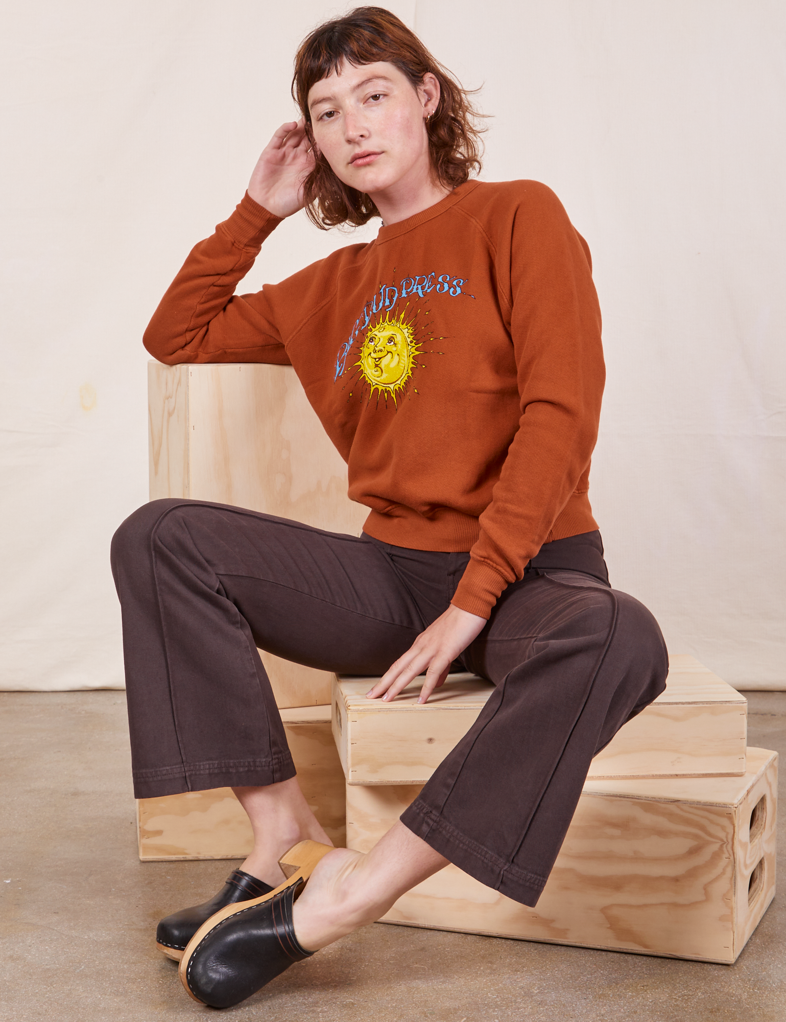 Alex is sitting on a wooden crate wearing Bill Ogden&#39;s Sun Baby Crew and espresso brown Western Pants