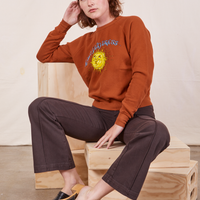 Alex is sitting on a wooden crate wearing Bill Ogden's Sun Baby Crew and espresso brown Western Pants