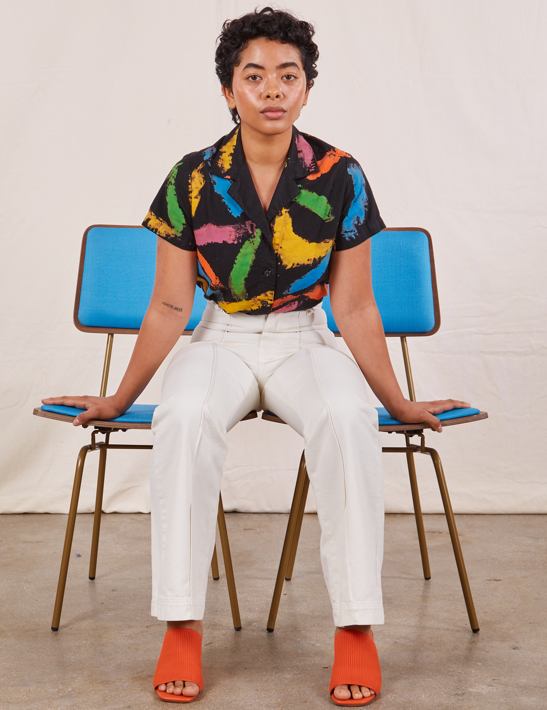 Mika is sitting in between two brass and blue chairs wearing Pantry Button Up in Paint Stroke and vintage off-white Western Pants.
