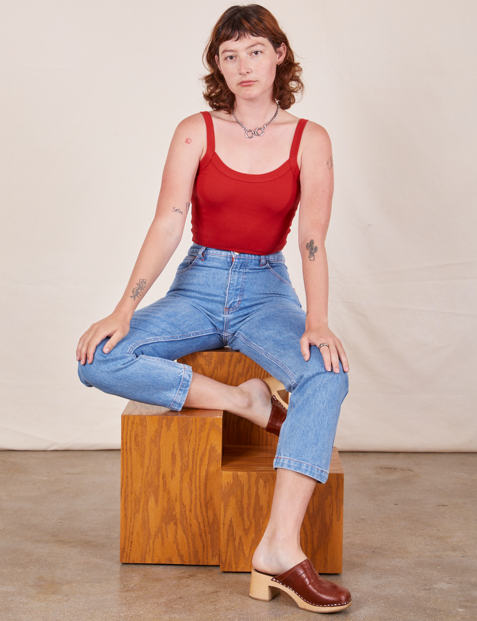 Alex is sitting on a wooden block wearing Cropped Cami in Mustang Red and light wash Frontier Jeans