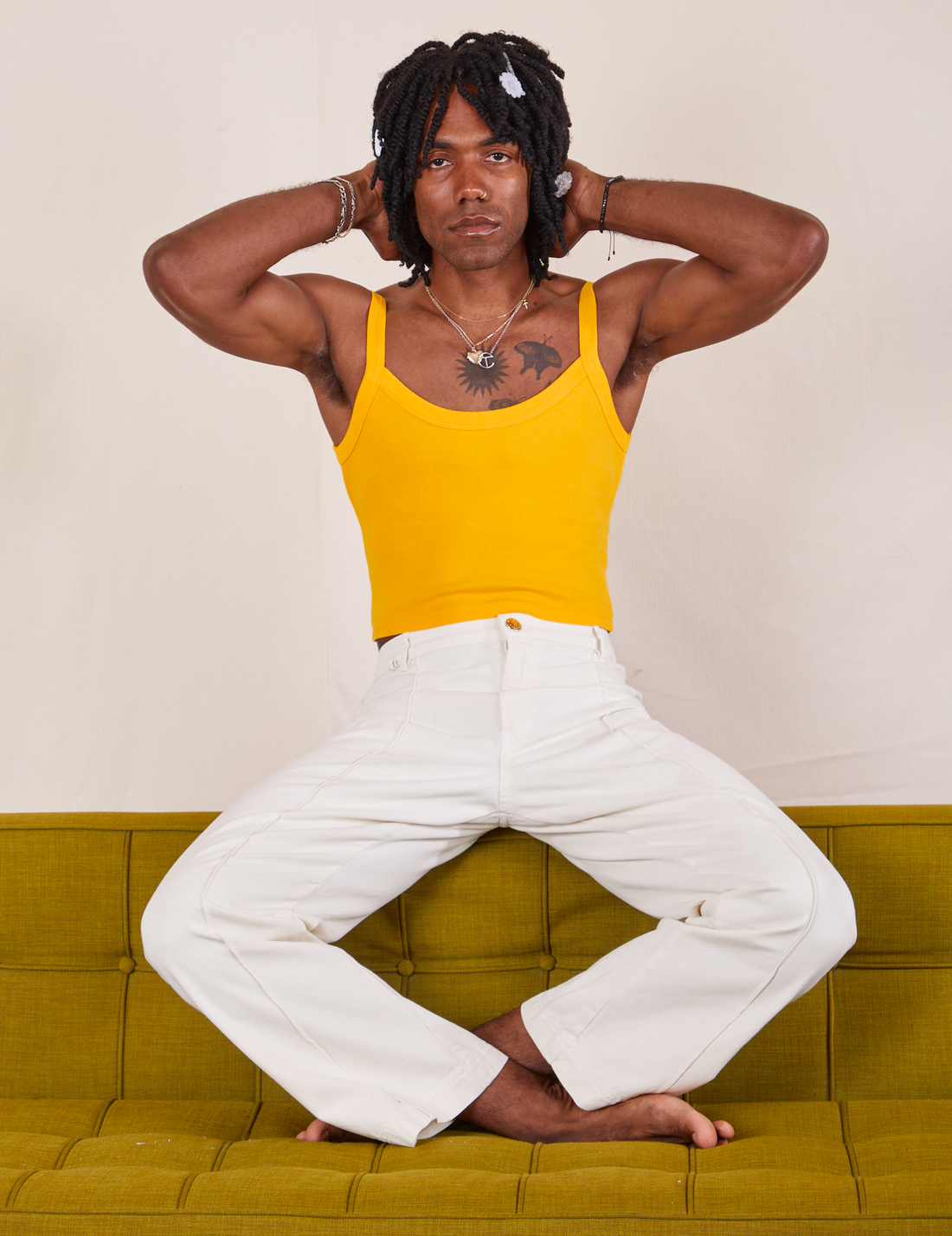 Jerrod is sitting on a olive green sofa wearing Cropped Cami in Sunshine Yellow and vintage off-white Western Pants