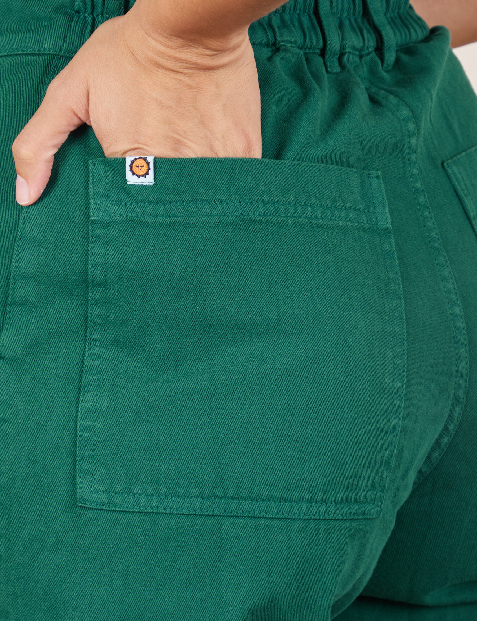 Back pocket close up of Short Sleeve Jumpsuit in Hunter Green. Tiara has her hand in the pocket.