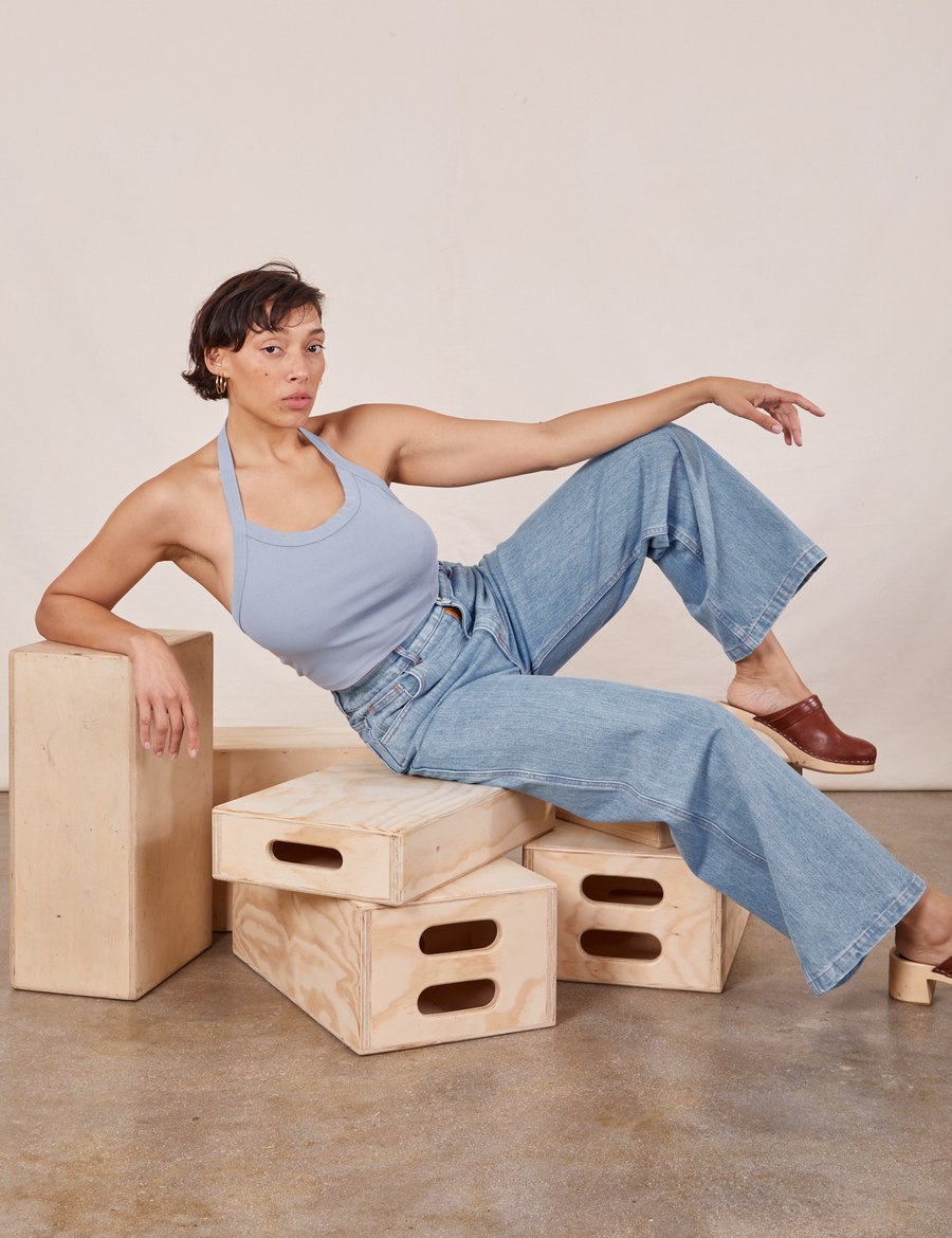 Tiara is sitting on a stack of wooden crate wearing Halter Top in Periwinkle and light wash Sailor Jeans