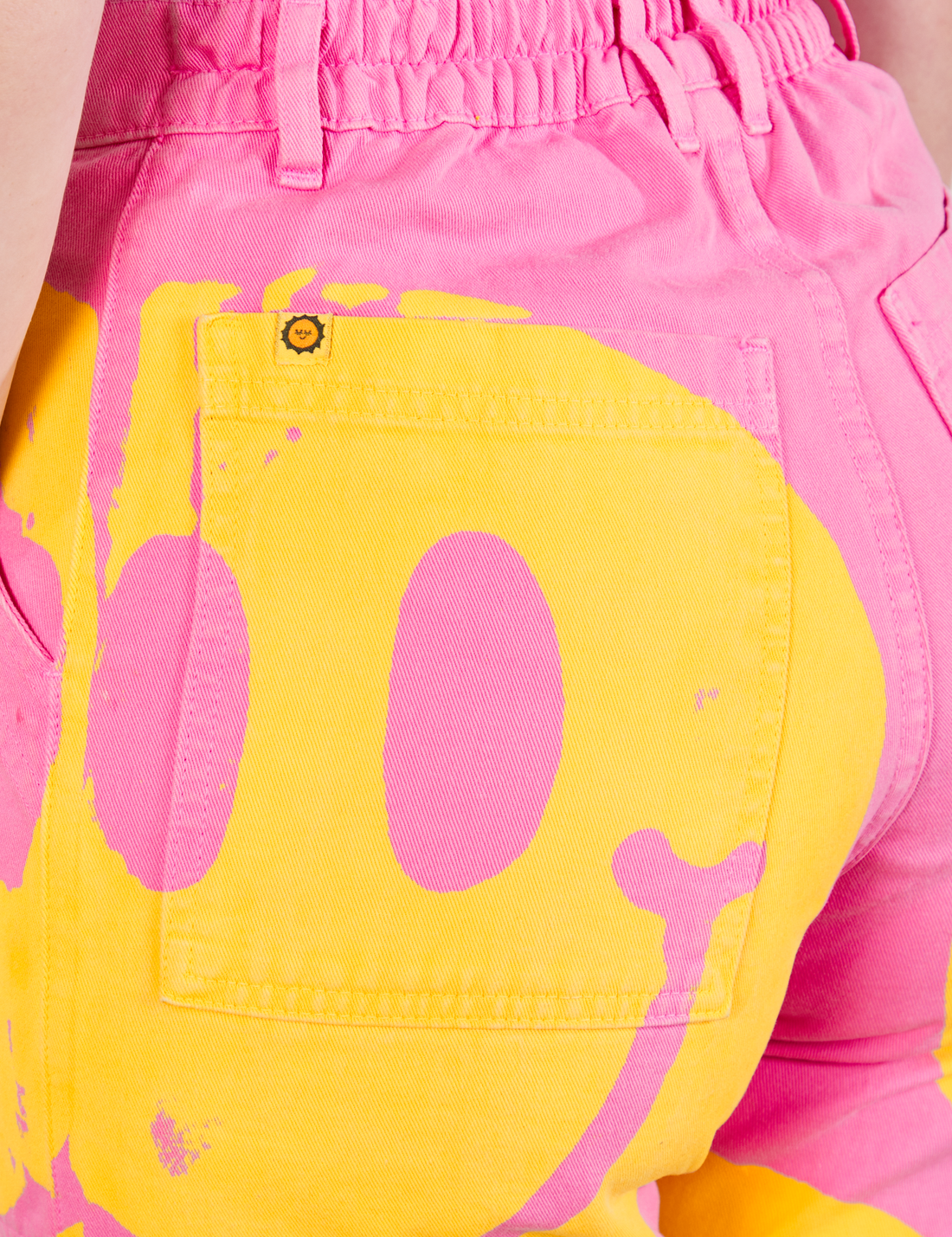 Back pocket close up of Icon Work Pants in Smilies worn by Alex. Bright Yellow paintstamped smiley face on a bubblegum pink background