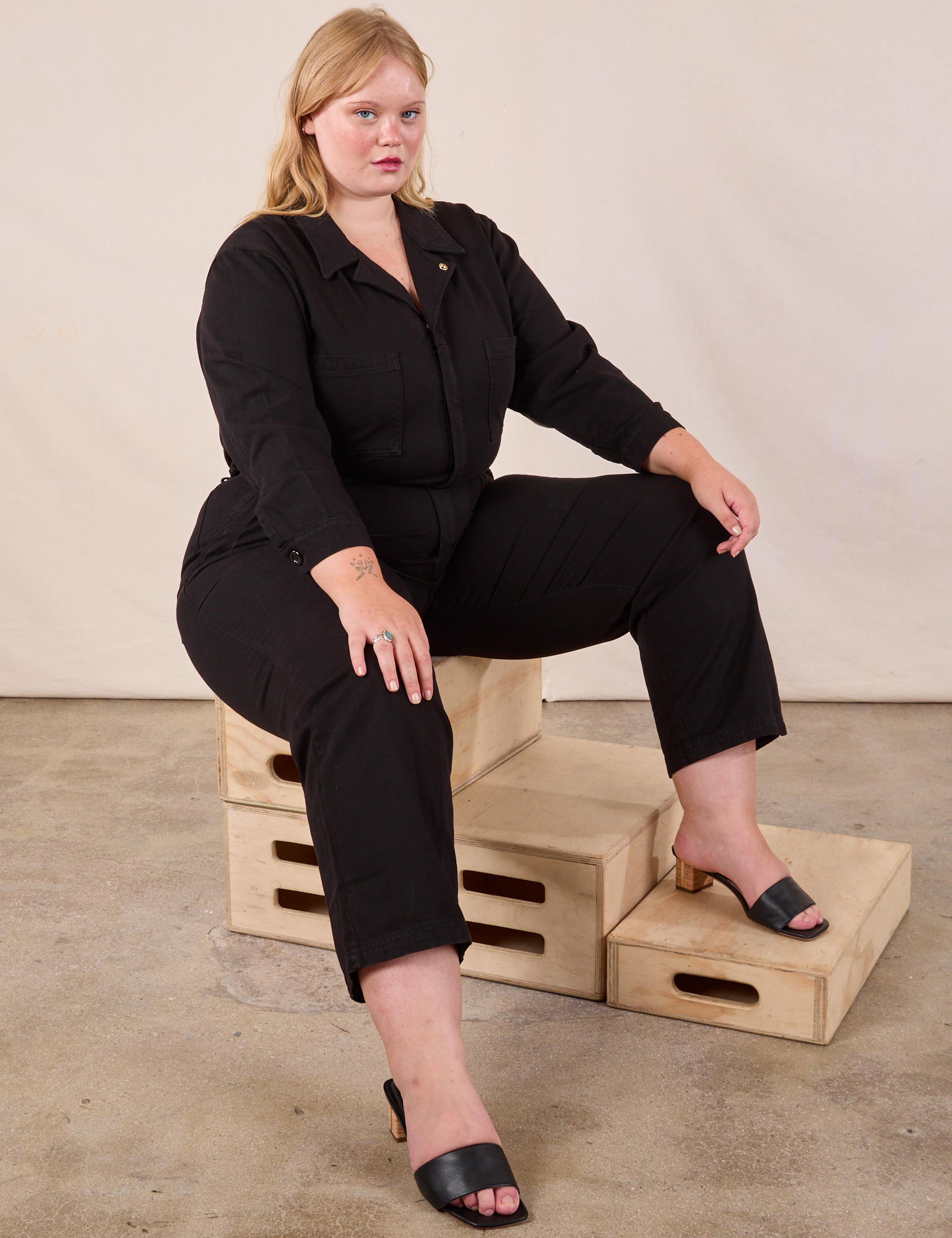 Juliet is wearing Long Sleeve Jumpsuit in Basic Black and sitting on a stack of crates.