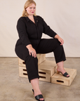 Juliet is wearing Long Sleeve Jumpsuit in Basic Black and sitting on a stack of crates.