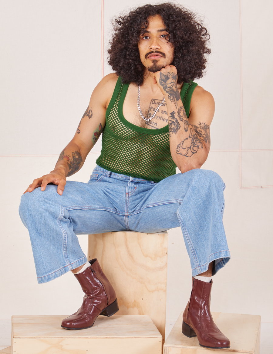 Jesse is sitting on a wooden crate wearing Mesh Tank Top in Lawn Green and light wash Sailor Jeans
