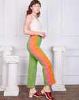 Side view of Hand-Painted Stripe Western Pants in Bright Olive and vintage off-white Tank Top worn by Alex. Pink and orange hand painted stripe down the side of the pant leg.