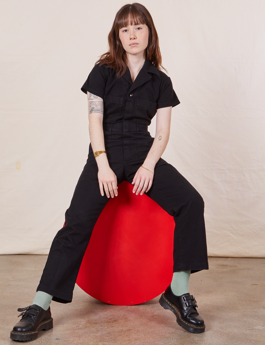Hana is sitting on a red circular stand wearing Petite Short Sleeve Jumpsuit in Basic Black