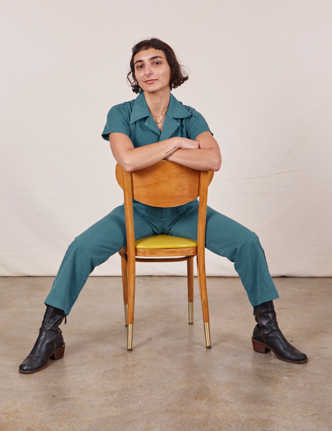 Soraya is sitting backwards on a wooden chair wearing Petite Short Sleeve Jumpsuit in Marine Blue