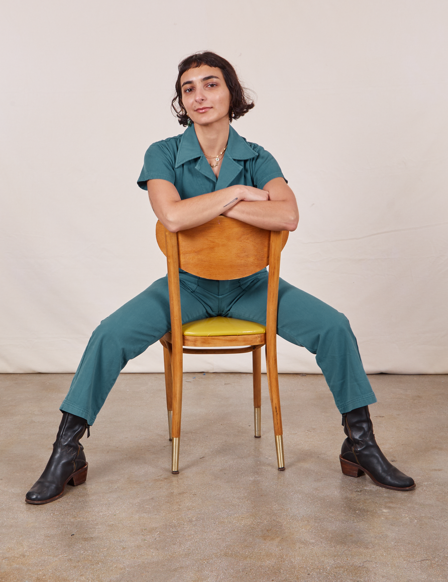Soraya is sitting backwards on a wooden chair wearing Petite Short Sleeve Jumpsuit in Marine Blue