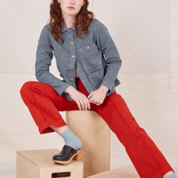 Alex is sitting on a wooden crate wearing Railroad Stripe Denim Work Jacket and paprika Western Pants