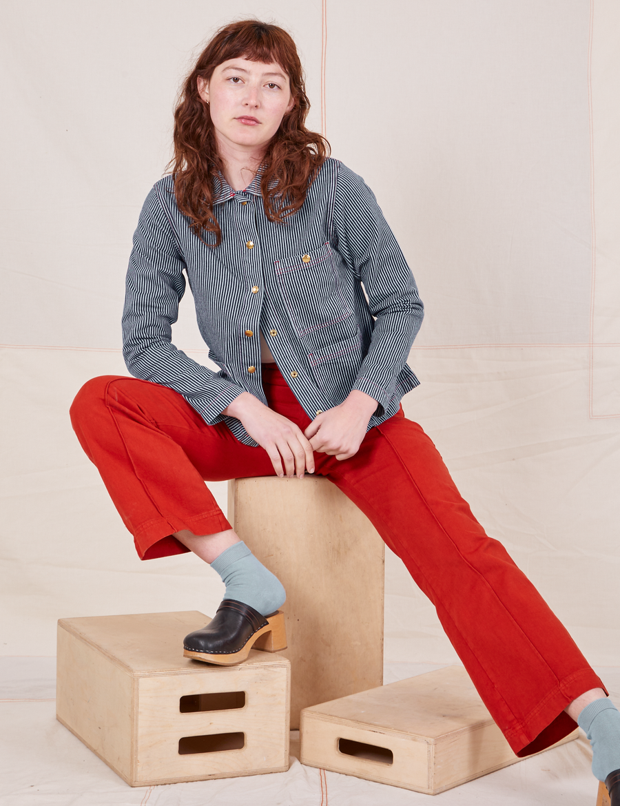 Alex is sitting on a wooden crate wearing Railroad Stripe Denim Work Jacket and paprika Western Pants