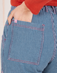 Back pocket close up of Railroad Stripe Denim Work Pants. Contrast red stitching and white and red sun baby logo tag on pocket. Tiara has her hand in the back pocket.