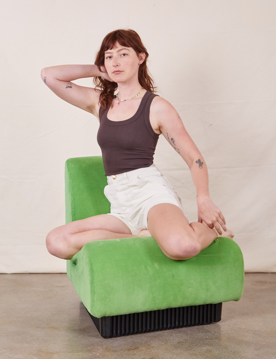 Alex is sitting on a green upholstered chair wearing  Classic Work Shorts in Vintage Off-White and espresso brown Tank Top