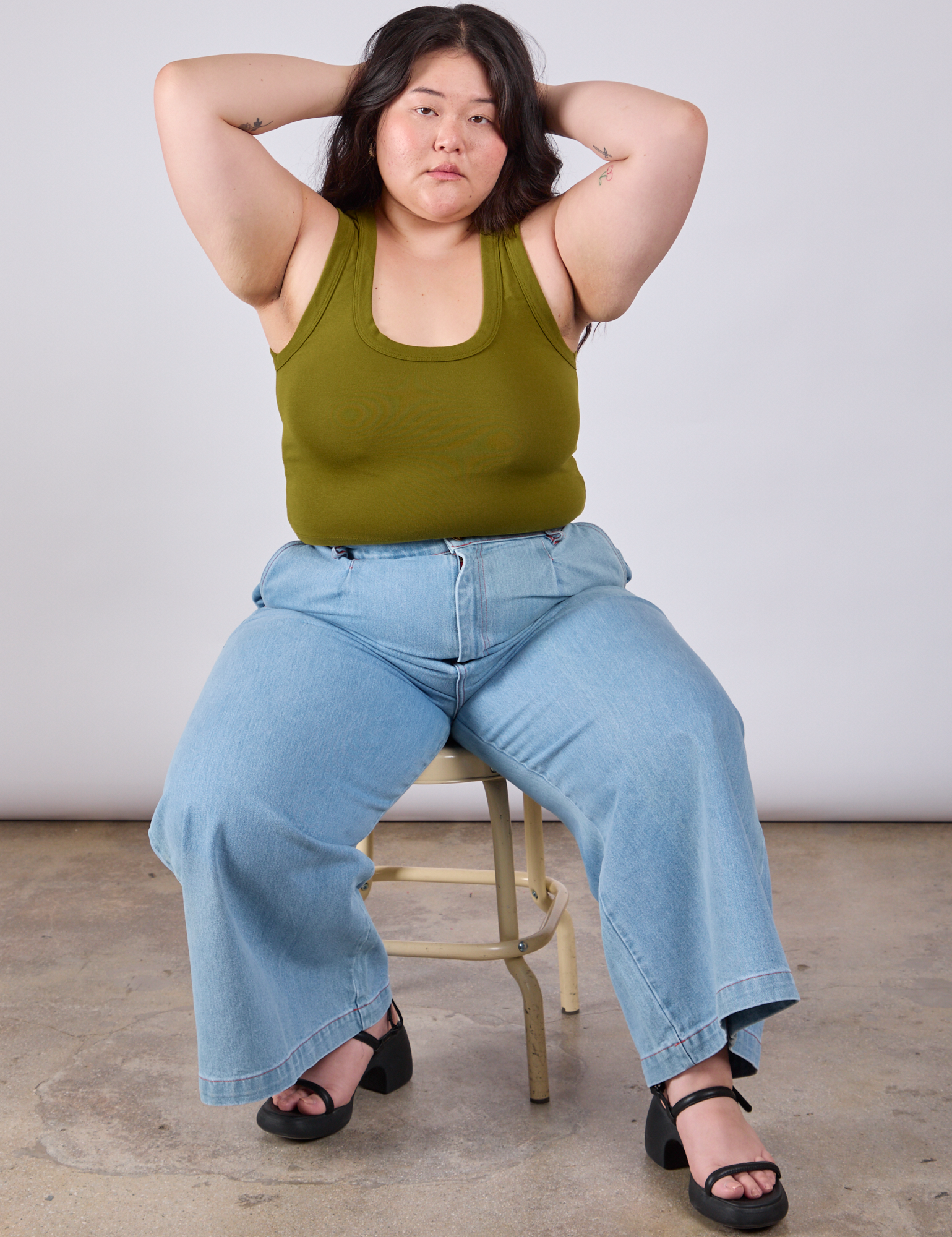 Ashley is sitting in a chair wearing Cropped Tank Top in Olive Green and light wash Indigo Wide Leg Trousers