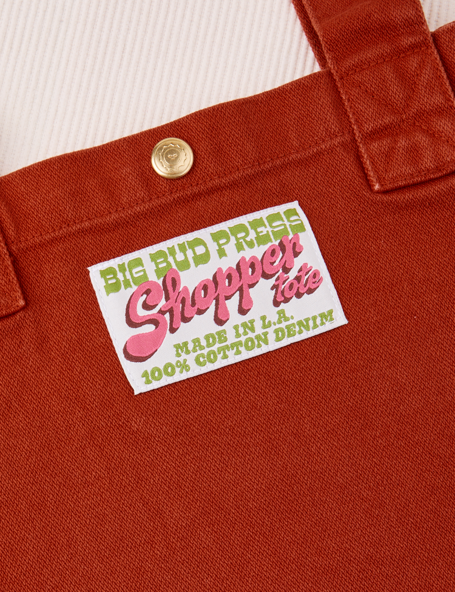 Close up of brass sun baby snap on paprika Shopper Tote Bag. Bag label with green and pink text that reads &quot;Big Bud Press, Shopper Tote, Made in L.A. 100% Cotton Denim&quot; on a white background