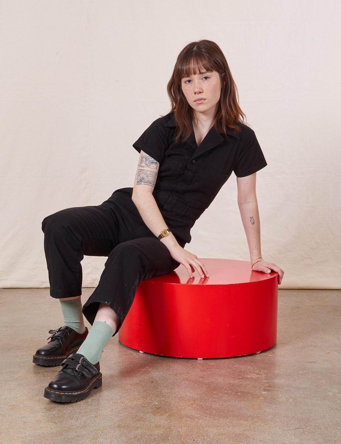 Hana is sitting on a red circular stand wearing Petite Short Sleeve Jumpsuit in Basic Black