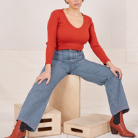 Tiara is sitting on a wooden crate wearing Railroad Stripe Denim Work Pants and a paprika Long Sleeve V-Neck Tee