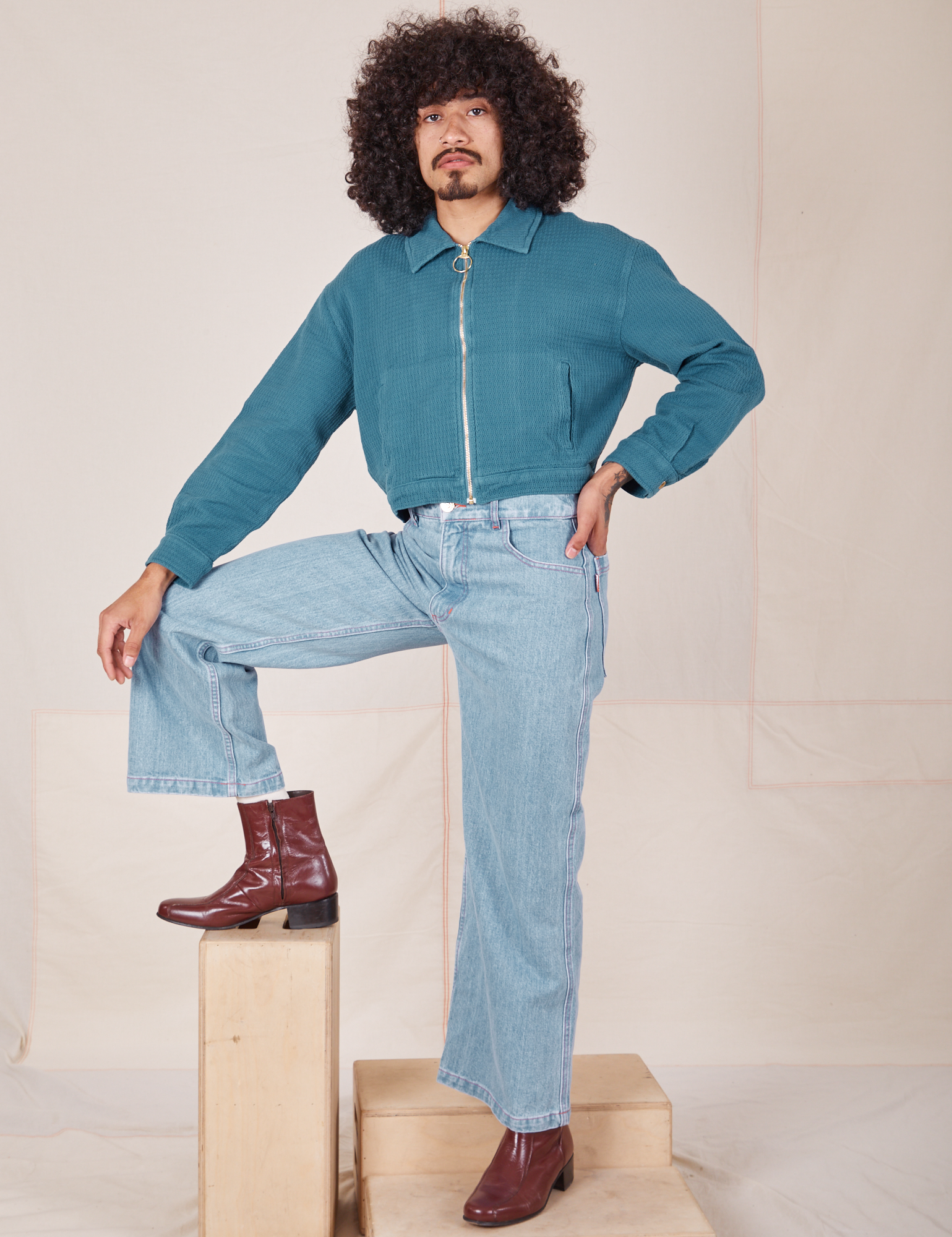 Jesse is standing on wooden crates wearing a zipped up Ricky Jacket in Marine Blue and light wash Frontier Jeans