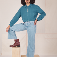Jesse is standing on wooden crates wearing a zipped up Ricky Jacket in Marine Blue and light wash Frontier Jeans