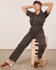 Tiara is sitting on a stack of wooden crates wearing Short Sleeve Jumpsuit in Espresso Brown.