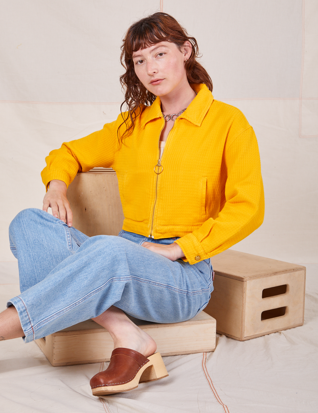 Alex is sitting on a wooden crate wearing the Ricky Jacket in Sunshine Yellow