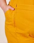Front pocket close up of Short Sleeve Jumpsuit in Mustard Yellow. Marielena has her hand in the pocket.