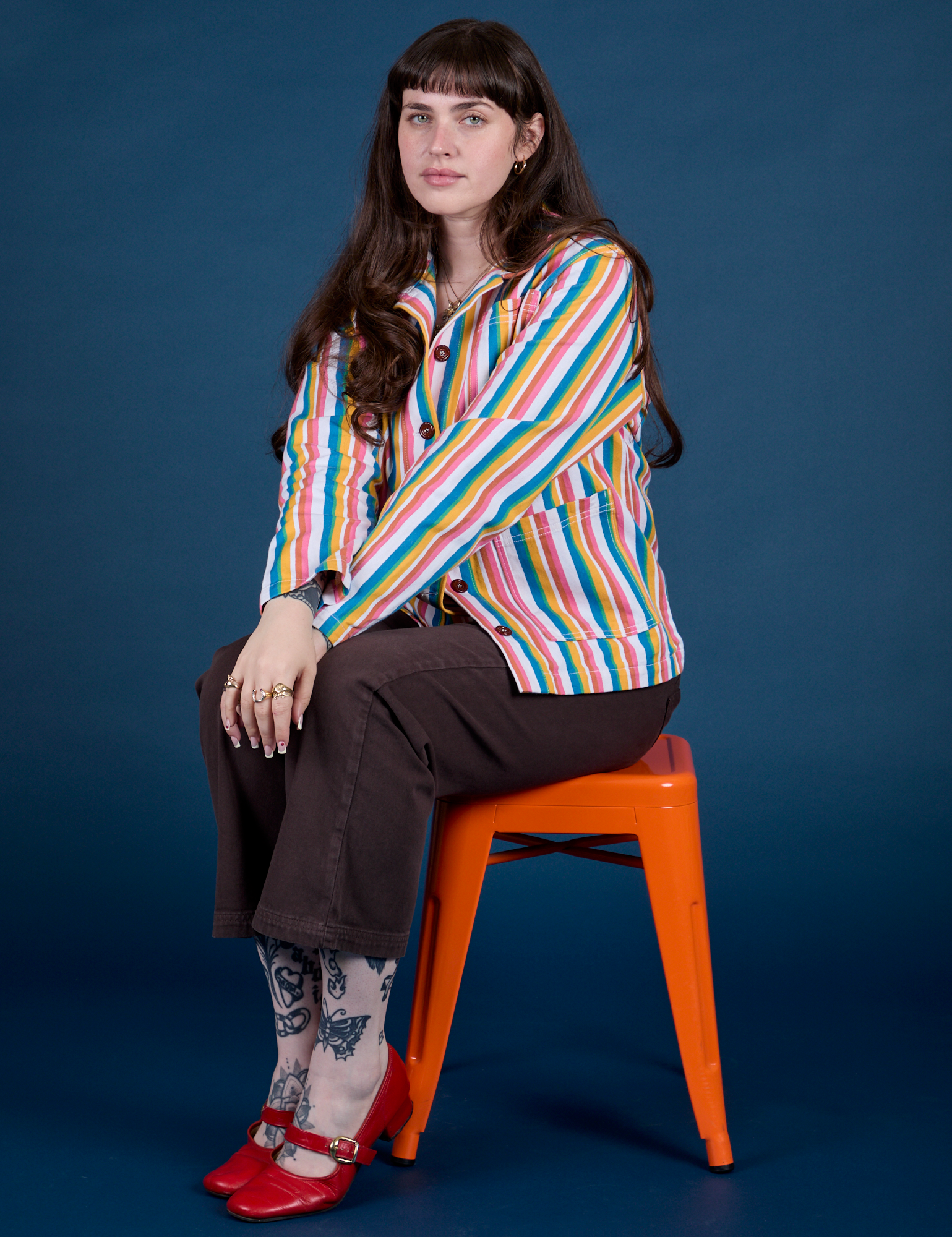 Sydney is sitting on a stool and is wearing Stripe Work Jacket in Rainbow Stripe paired with espresso brown Work Pants