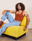 Jesse is sitting in a yellow upholstered chair. They are wearing Cropped Tank Top in Burnt Terracotta and light wash Sailor Jeans