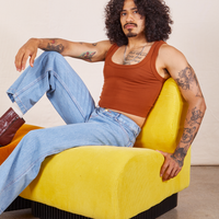 Jesse is sitting in a yellow upholstered chair. They are wearing Cropped Tank Top in Burnt Terracotta and light wash Sailor Jeans