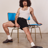 Jesse is sitting in a vintage brass and blue chair wearing Trouser Shorts in Basic Black and a vintage off-white Tank Top