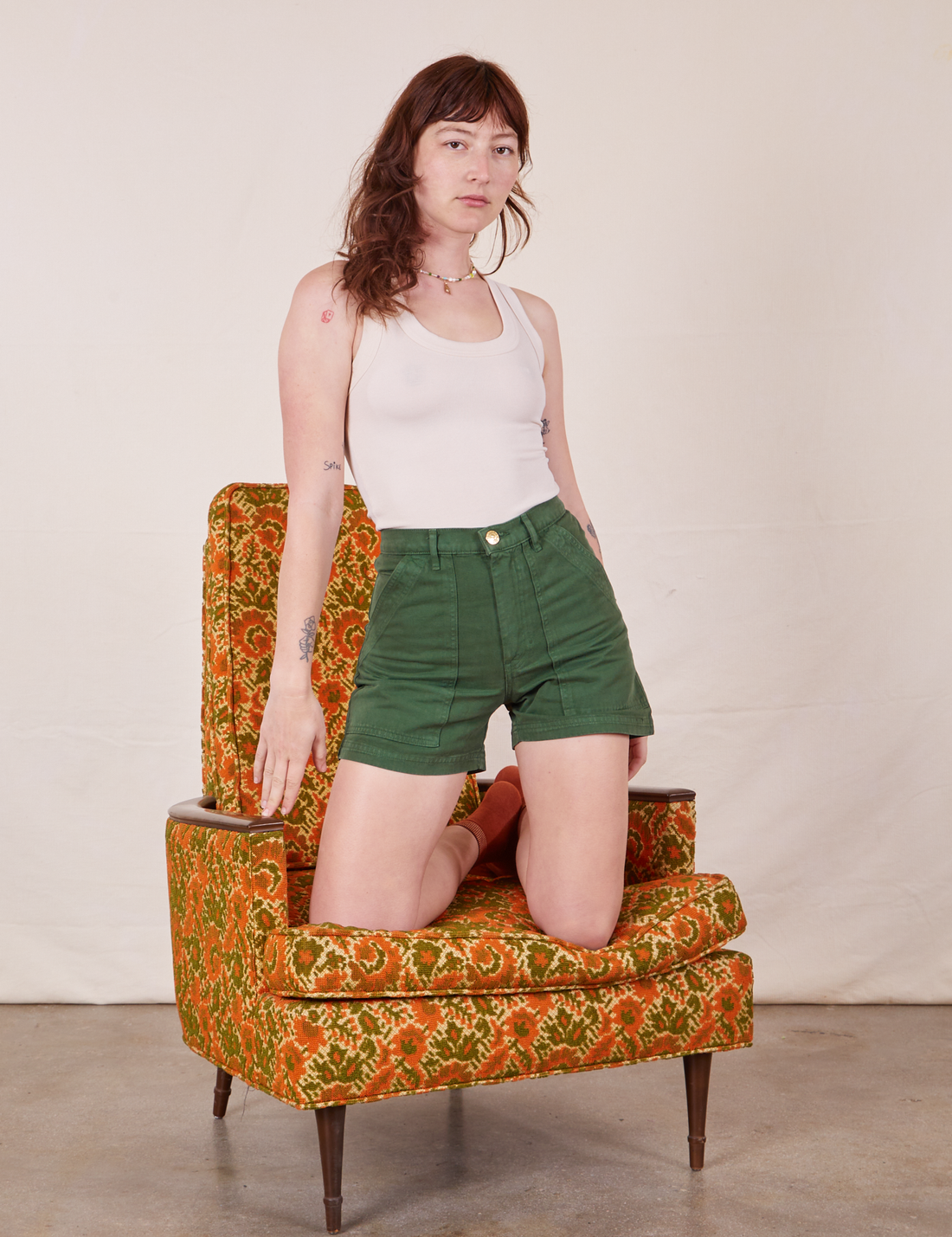 Alex is kneeling on a vintage floral upholstered chair wearing Classic Work Shorts in Dark Emerald Green and a vintage off-white Tank Top