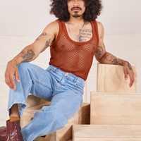 Jesse is sitting on a wooden crate wearing Mesh Tank Top in Burnt Terracotta and light wash Sailor Jeans