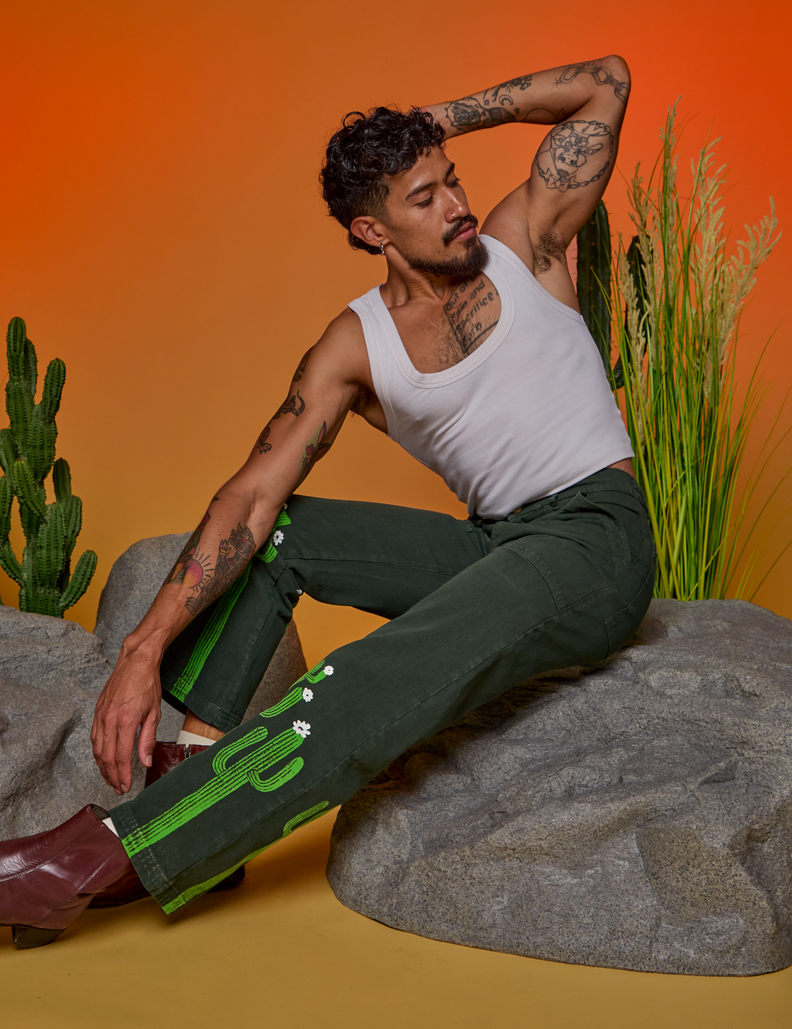 Jesse is sitting on a boulder. They are wearing Paintstamped Work Pants in Cactus and a Cropped Tank in Vintage Tee White