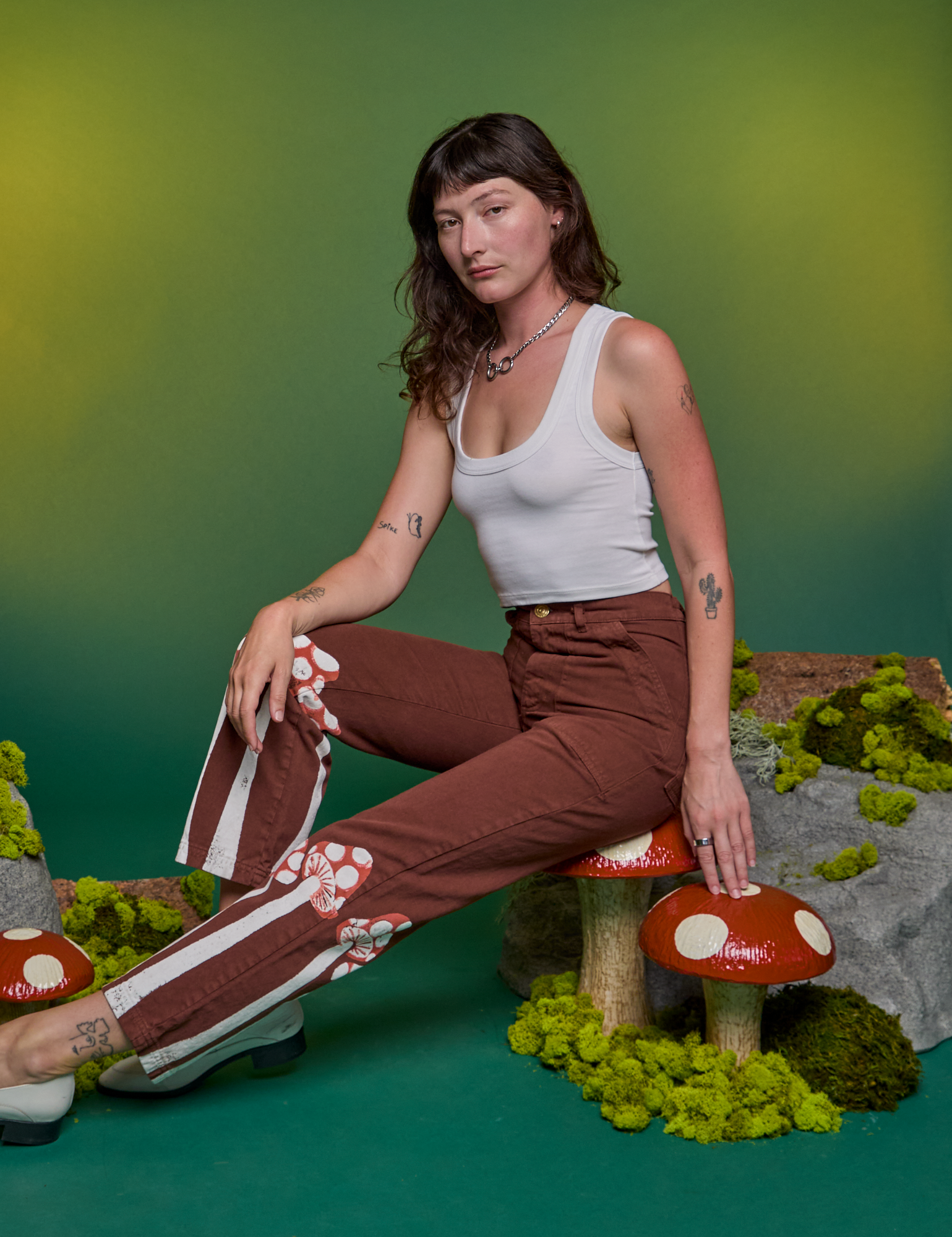 Alex is sitting on a mushroom and wearing Paintstamped Work Pants in Mushroom paired with a Cropped Tank in Vintage Tee White