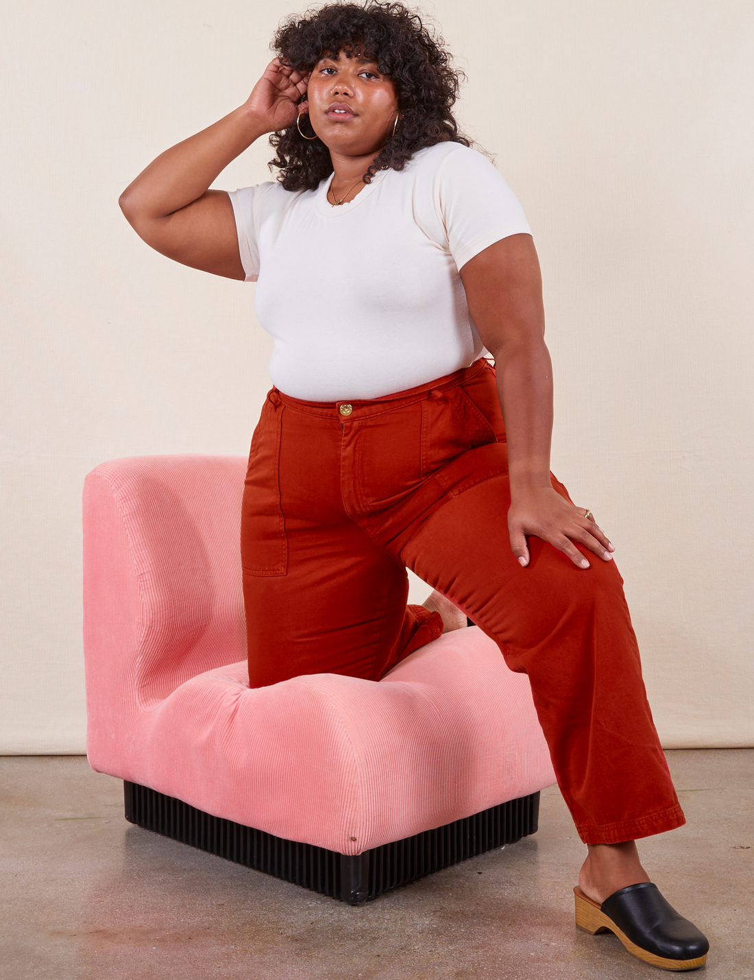 Work Pants in Paprika on Morgan wearing a vintage off-white Baby Tee. Morgan has one knee on a pink upholstered chair and the other leg on the ground.