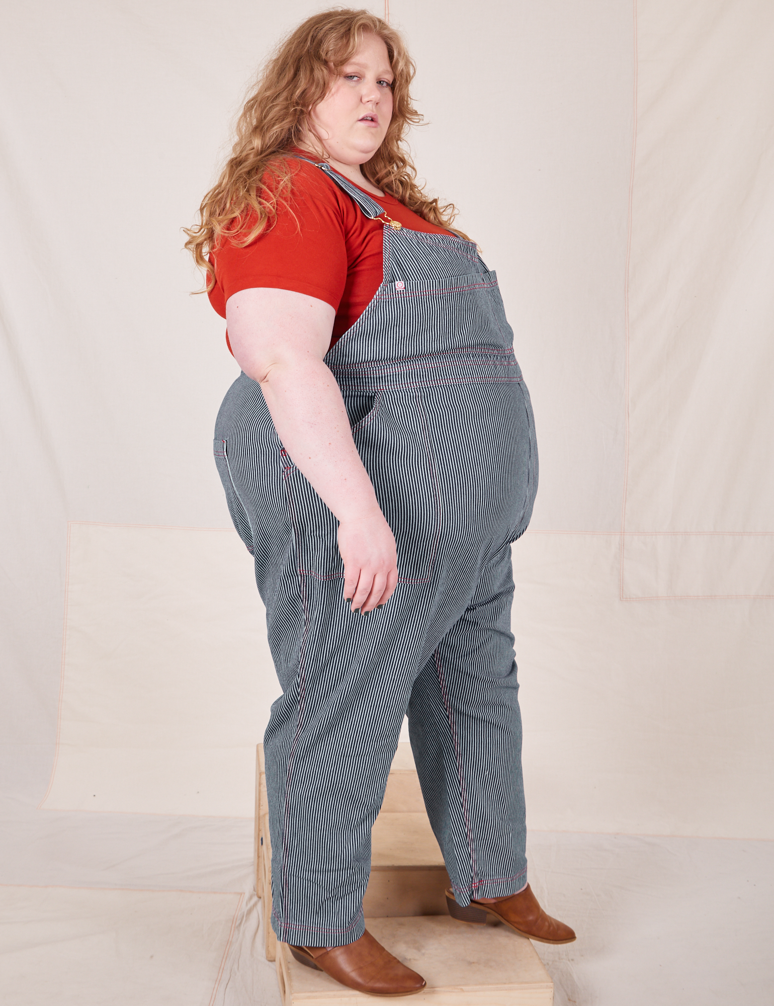 Side view of Railroad Stripe Denim Original Overalls and paprika Baby Tee worn by Catie