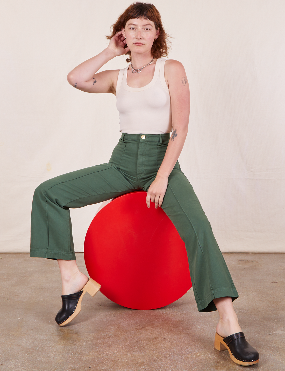 Alex is sitting on a circular red stand wearing Western Pants in Dark Green Emerald and a vintage off-white Tank Top