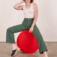 Alex is sitting on a circular red stand wearing Western Pants in Dark Green Emerald and a vintage off-white Tank Top