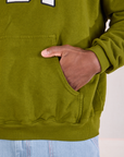 Collegiate Hoodie in Summer Olive kangaroo pocket close up. Issac has his hand in the pocket.