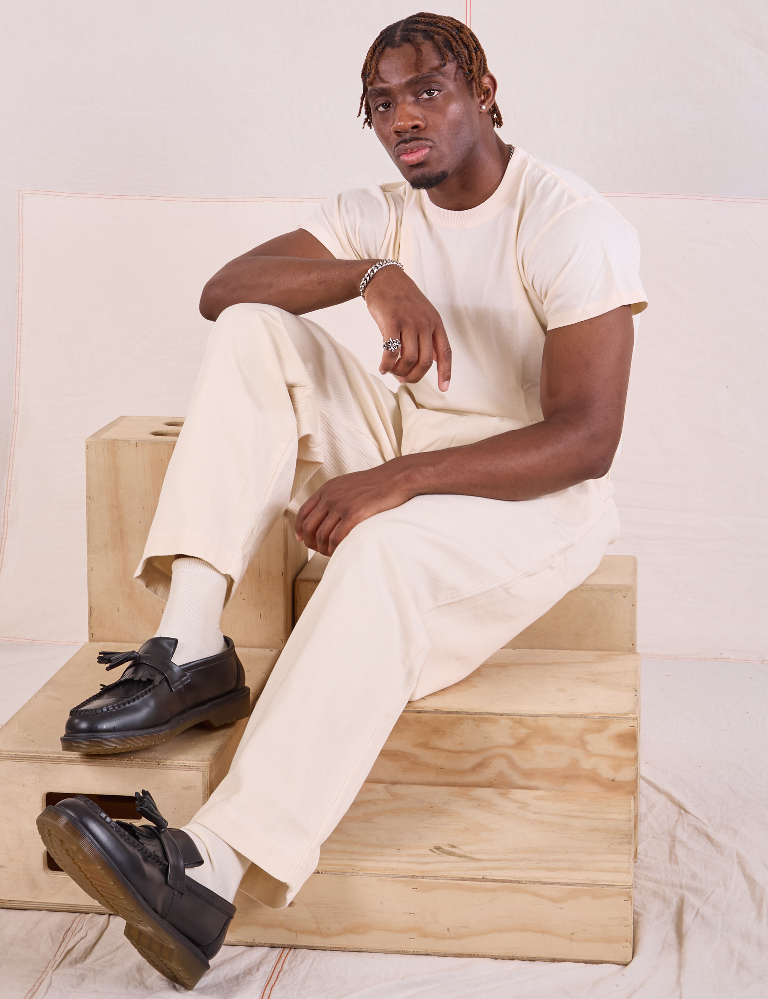 Issac is sitting on a wooden crate. He is wearing Heritage Trousers in Natural and Organic Vintage Tee in vintage tee off-white