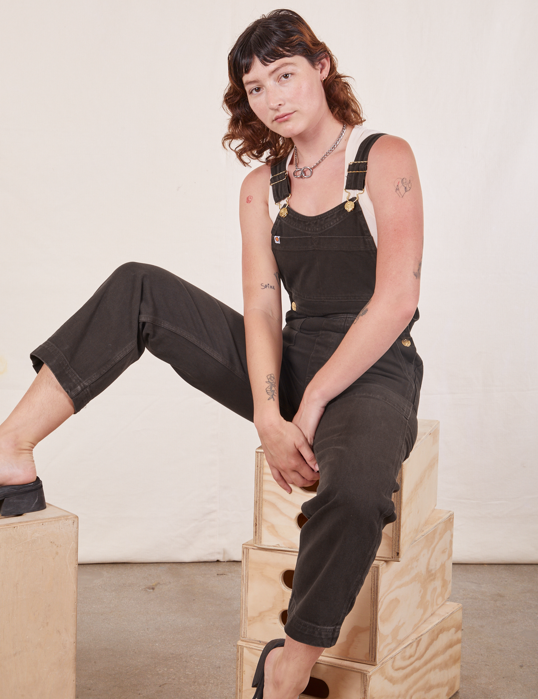 Alex is sitting on a stack of wooden crates wearing Original Overalls in Mono Espresso.