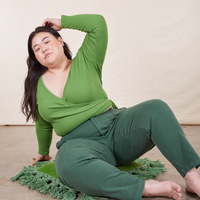 Ashley is sitting on a green cushion wearing Pencil Pants in Dark Emerald Green paired with a bright olive Wrap Top